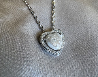 May include: A silver-toned necklace featuring a heart-shaped pendant with a textured, off-white center stone. The pendant is framed by a border of small, clear, sparkling stones and hangs from a delicate, matching silver-toned chain.