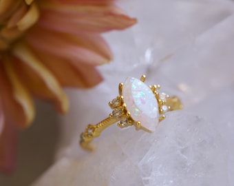 May include: A gold ring featuring a central marquise-cut white opal with iridescent flashes, flanked by small clear stones on each side. The thin gold band has a textured, branch-like appearance and is set with additional small clear stones.