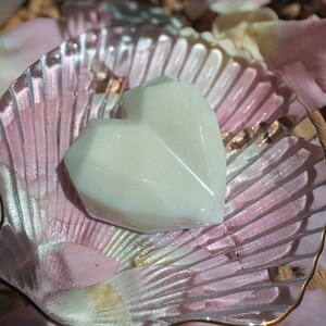 May include: A heart-shaped, off-white soap sits on a clear, shell-shaped dish with a gold rim. The dish is surrounded by pink and white rose petals, creating a soft, romantic aesthetic. The soap has a faceted design, adding visual interest.