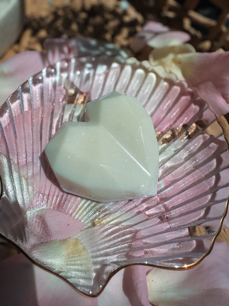May include: A heart-shaped, off-white soap sits on a clear, shell-shaped dish with a gold rim. The dish is surrounded by pink and white rose petals, creating a soft, romantic aesthetic. The soap has a faceted design, adding visual interest.