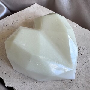 May include: A pale green, heart-shaped object with a faceted design. The object has a subtle shimmer and sits on a textured, light-colored surface. The background features a draped, silvery fabric.