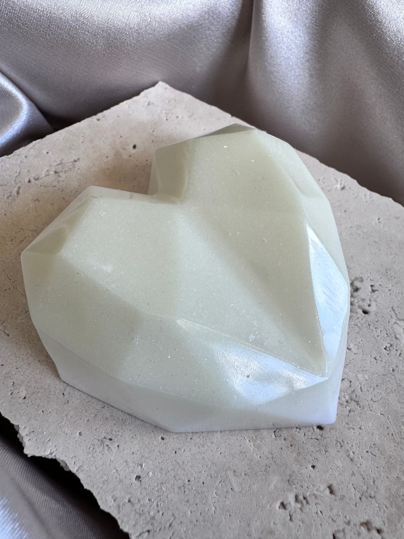 May include: A pale green, heart-shaped object with a faceted design. The object has a subtle shimmer and sits on a textured, light-colored surface. The background features a draped, silvery fabric.