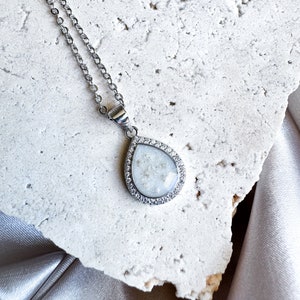 Peut inclure: Un collier en chaîne argent avec un pendentif en forme de larme. Le pendentif est rempli de paillettes blanches et a une bordure transparente et scintillante.