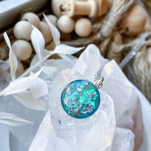 May include: A round pendant necklace with a silver chain. The pendant features a teal and silver design, with a mix of teal and silver flakes. The necklace is displayed on a clear, crystalline surface.