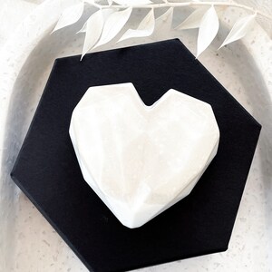 May include: A white, geometric heart-shaped object rests on a black, hexagonal base. The heart has a marbled appearance and is set against a neutral background. Decorative white leaves are placed above the heart.