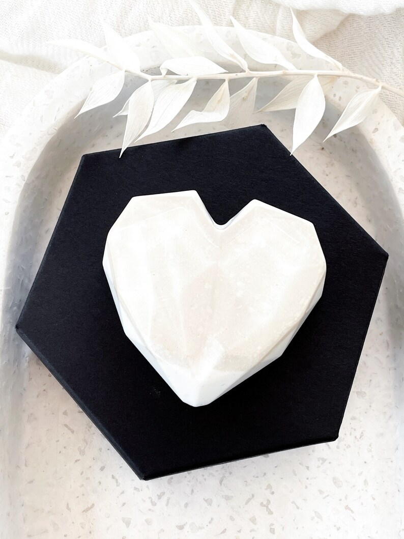 May include: A white, geometric heart-shaped object rests on a black, hexagonal base. The heart has a marbled appearance and is set against a neutral background. Decorative white leaves are placed above the heart.
