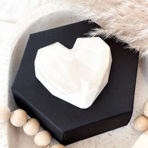 May include: A white, geometric heart-shaped object rests on a black, hexagonal box. The heart has a faceted design. The background includes decorative elements like dried pampas grass and wooden beads, creating a minimalist aesthetic.
