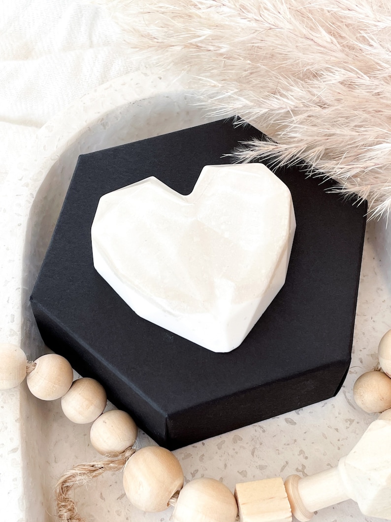 May include: A white, geometric heart-shaped object rests on a black, hexagonal box. The heart has a faceted design. The background includes decorative elements like dried pampas grass and wooden beads, creating a minimalist aesthetic.