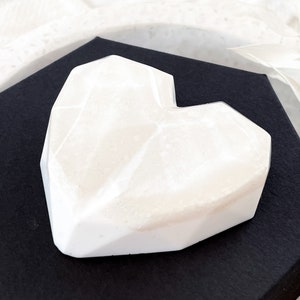 May include: A white, geometric heart-shaped object with a faceted design. The heart is resting on a black surface, creating a contrast. The object appears to be made of a solid, opaque material.