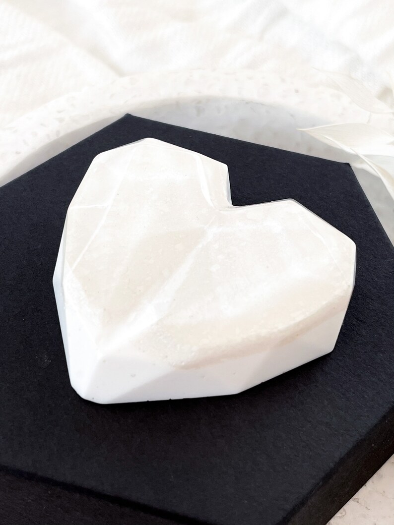 May include: A white, geometric heart-shaped object with a faceted design. The heart is resting on a black surface, creating a contrast. The object appears to be made of a solid, opaque material.