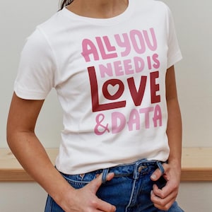 Pode incluir: Camiseta branca com a frase "ALL YOU NEED IS LOVE & DATA" em letras rosa e bordô. A camiseta tem gola redonda e mangas curtas. A pessoa está usando jeans azul.
