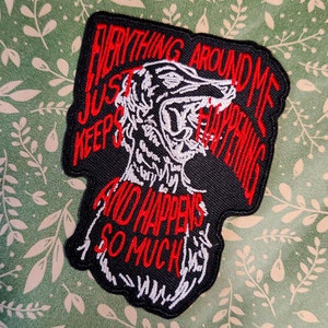 May include: Black and white embroidered patch with a red outline. The patch features a stylised illustration of a wolf's head with the text "Everything around me just keeps happening and happens so much".