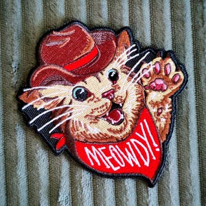 May include: An embroidered patch featuring a tan tabby cat wearing a brown cowboy hat and a red bandana. The cat has one paw raised in a wave, and the red bandana displays the white text MEOWDY! The design is outlined in black thread.