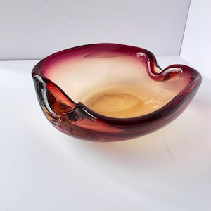 May include: A vintage glass ashtray with a unique, organic shape. The ashtray features a gradient of colors, transitioning from a deep burgundy rim to a warm amber center. The glass has a glossy finish, reflecting light and adding to its visual appeal.