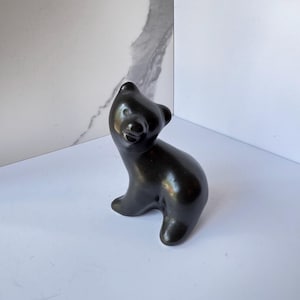 May include: A smooth, polished black bear figurine in a seated position with its head tilted upward. The sculpture features a rounded body, small ears, and a defined snout, showcasing a solid dark finish with subtle light reflections on its surface.