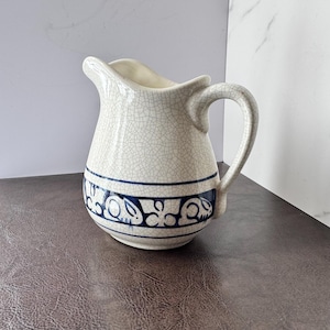 May include: Cream-colored ceramic pitcher with a crackle glaze. A dark blue band with a repeating floral design encircles the base. The pitcher has a curved handle and a pouring spout.