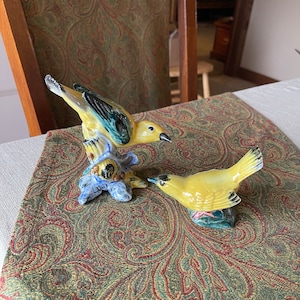 May include: Two ceramic bird figurines. One yellow bird with black and teal accents is perched on a blue and brown tree branch. The other yellow bird is on a small green base. Both have black beaks.