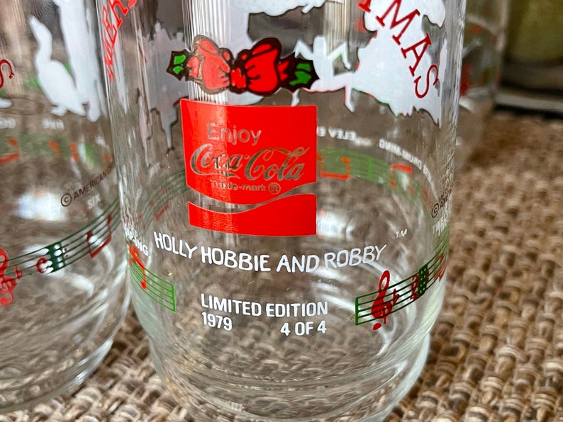 May include: Clear glass Coca-Cola Holly Hobbie and Robby Christmas glasses. Each glass features a red Coca-Cola logo, musical notes, and the text "Limited Edition 1979 4 of 4". The glasses have a festive design.