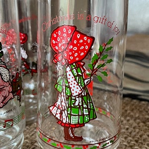 May include: Clear glass Holly Hobbie Christmas glasses. Each glass features a red-bonneted girl holding holly, with the text "Christmas is a gift of joy." The glasses have a red and green holly berry design around the base.