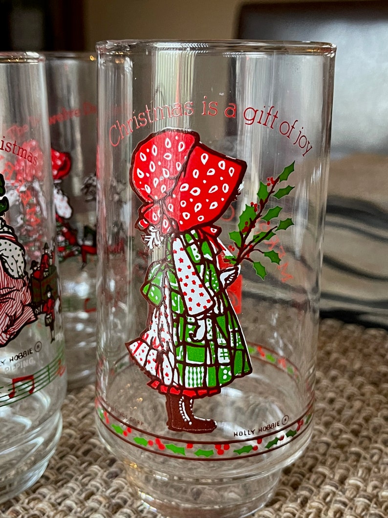 May include: Clear glass Holly Hobbie Christmas glasses. Each glass features a red-bonneted girl holding holly, with the text "Christmas is a gift of joy." The glasses have a red and green holly berry design around the base.