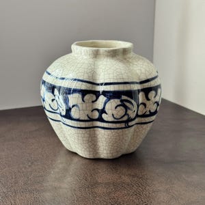May include: A cream-colored ceramic vase with a crackle glaze finish. The vase has a rounded shape with a fluted body and a dark blue decorative band featuring floral motifs. The vase is suitable for displaying flowers or as a decorative accent.
