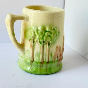 May include: Cream-colored ceramic mug with a handle, featuring a green and brown relief design of trees and foliage. The base and the area around the trees are green. The mug has a slightly textured surface.