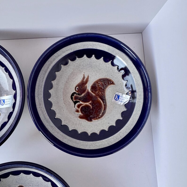 May include: Ceramic plate with a dark blue rim and a light gray center. The center features a brown squirrel design. The plate has a scalloped edge and a dark blue border. A sticker is visible on the plate.