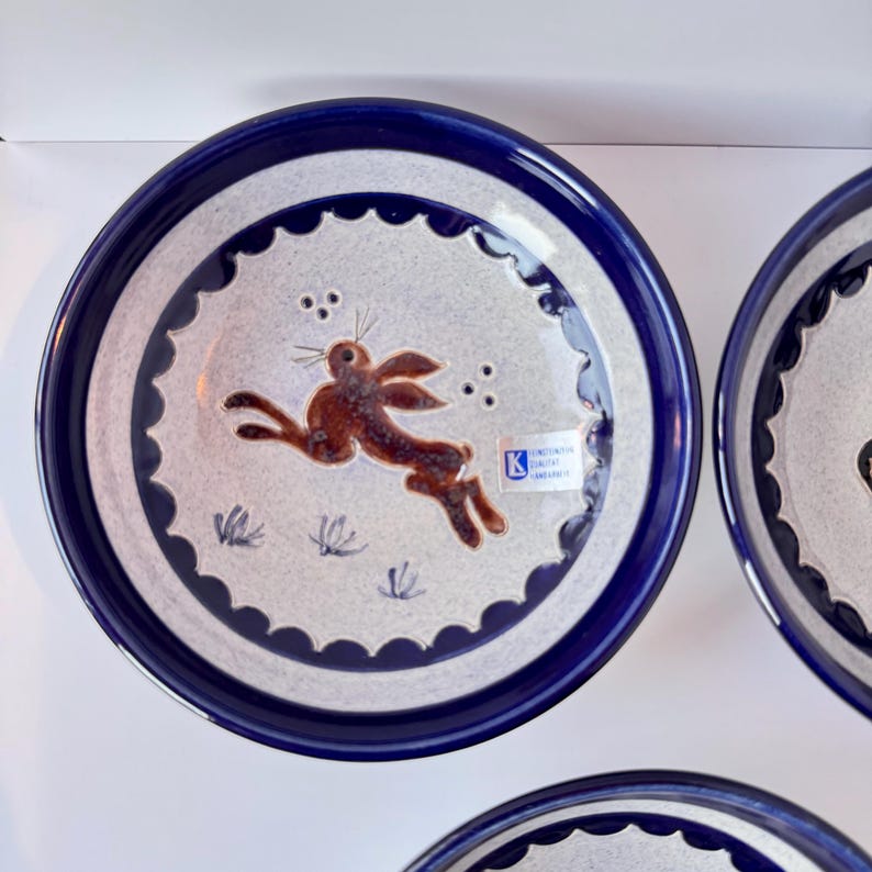 May include: Ceramic plate with a dark blue rim and a light gray center. The center features a brown rabbit leaping across the plate, with small grass and dot details. A sticker is visible on the plate.