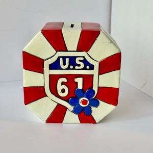 May include: A ceramic piggy bank in the shape of a cube, featuring a red, white, and blue color scheme. The front displays a U.S. 61 road sign design with a blue flower accent. The top has a slot for inserting coins.