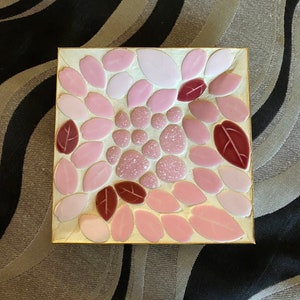 May include: Square mosaic tile art featuring a floral design. The mosaic is composed of pink, light pink, and burgundy leaf-shaped tiles and speckled round tiles. The tiles are set on a gold-colored background and framed with a gold border.