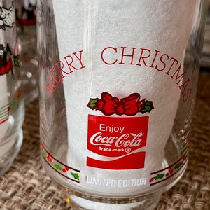 May include: Clear glass Coca-Cola Christmas glass with red text that reads "MERRY CHRISTMAS" and "Enjoy Coca-Cola". The glass has a red bow and holly design and the words "LIMITED EDITION".