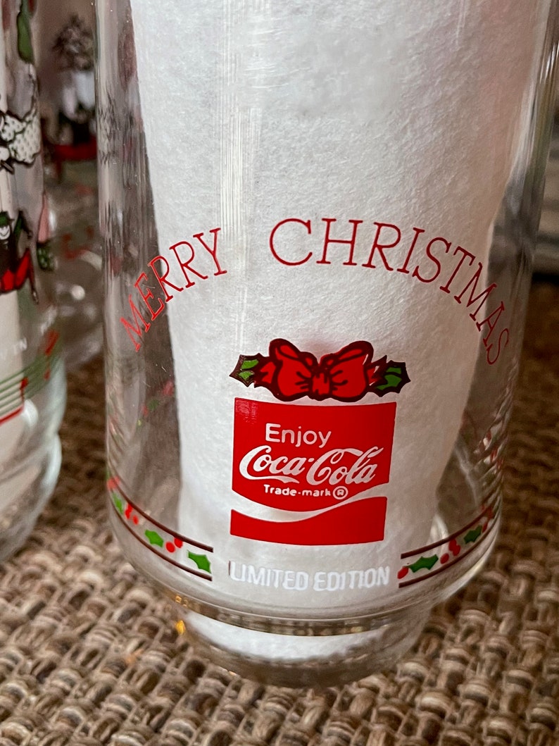 May include: Clear glass Coca-Cola Christmas glass with red text that reads "MERRY CHRISTMAS" and "Enjoy Coca-Cola". The glass has a red bow and holly design and the words "LIMITED EDITION".