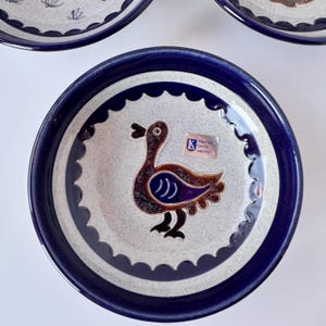 May include: A set of ceramic plates with a duck design. The plates have a blue rim and a white center with a repeating scalloped pattern. The duck is brown with blue and purple accents. A sticker is visible on the plate.