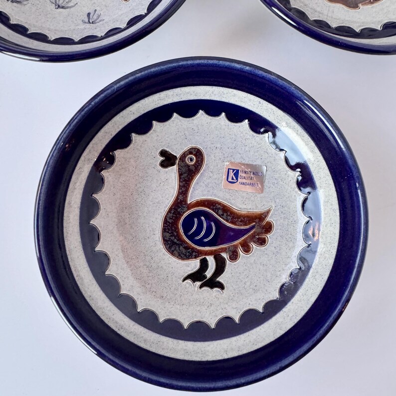 May include: A set of ceramic plates with a duck design. The plates have a blue rim and a white center with a repeating scalloped pattern. The duck is brown with blue and purple accents. A sticker is visible on the plate.
