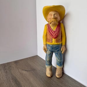 May include: A carved wooden figurine of a cowboy. The cowboy wears a yellow hat, shirt, and boots, a red scarf, a brown vest, and blue jeans. The figurine has a large mustache and is standing upright.