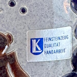 May include: Close-up of a ceramic piece with a label. The label has the letter "K" inside a blue square, and the words "FEINSTEINZEUG QUALITAT HANDARBEIT" are printed below it. The background is a light gray color.