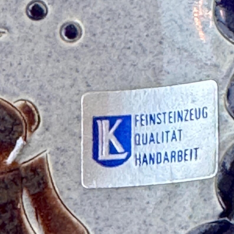 May include: Close-up of a ceramic piece with a label. The label has the letter "K" inside a blue square, and the words "FEINSTEINZEUG QUALITAT HANDARBEIT" are printed below it. The background is a light gray color.
