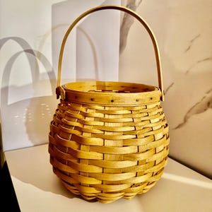 May include: A woven basket with a rounded base and a handle. The basket is made of light brown, interwoven strips of wood. The handle arches over the top, attached to the rim. The basket is empty and sits on a white surface.
