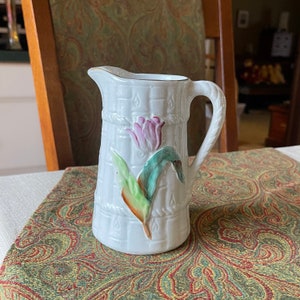 May include: White ceramic creamer with a floral design. The creamer features a pink tulip with green leaves on a white background. The creamer has a handle and a spout.