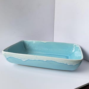 May include: A rectangular, light blue ceramic baking dish with a white, wavy trim. The dish has rounded corners and a smooth interior. The exterior has a glossy finish. The dish is empty and sits on a white surface.