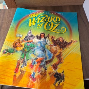 May include: A colorful promotional poster for "The Wizard of Oz on Ice" featuring Dorothy, the Tin Man, Scarecrow, and the Cowardly Lion. The poster has a rainbow and the title in large, yellow, stylized letters. A small black dog is also visible.