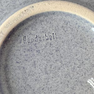 May include: Close-up of a speckled, light blue ceramic bowl with a cream-colored rim. The word "Handarbeit" is stamped in the center of the bowl. The bowl has a textured surface.