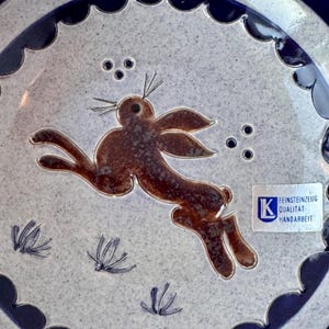 May include: A ceramic plate with a brown rabbit design jumping across the center. The plate has a light blue speckled background with a dark blue scalloped edge. The plate also has a small white sticker with German text.