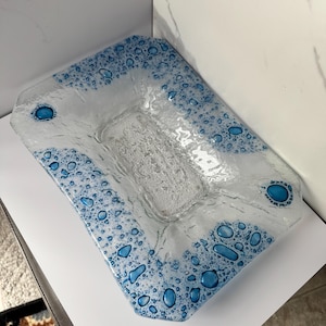 May include: A rectangular glass serving dish with a textured surface and a blue bubble design around the edges. The center of the dish has a raised, textured pattern. The corners feature larger blue circles.