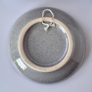 May include: A gray ceramic plate with a speckled texture, featuring a cream-colored rim and a white string for hanging. The plate has the text "Handarbeit" and "West Germany" on the back.