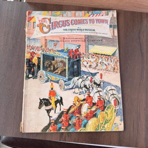 May include: Vintage book cover titled "The Circus Comes to Town" featuring a circus parade scene. The cover shows a lion cage on wheels, a marching band, and a crowd of people. The book is a publication of The Circus World Museum.