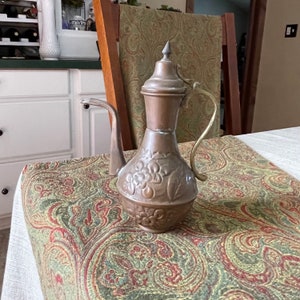 May include: A copper-colored, vintage-style pitcher with a long spout and a handle. The pitcher features floral embossed detailing around the base and a pointed lid. The handle and spout are a lighter brass color. The pitcher is sitting on a paisley patterned tablecloth.