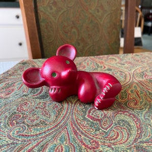 May include: A red ceramic mouse figurine with green eyes and a toothy grin. The mouse is lying down, with its paws and ears visible. The figurine is on a patterned tablecloth.
