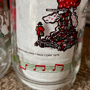 May include: Clear glass with a festive illustration of a person playing a flute, wearing a red and green patterned dress and hat. The glass has the text "Twelve Drummers Drumming" and "American Greetings Corp. 1979".