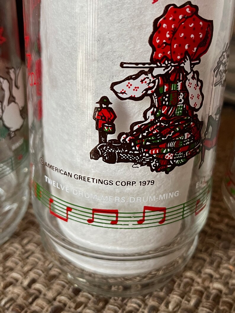 May include: Clear glass with a festive illustration of a person playing a flute, wearing a red and green patterned dress and hat. The glass has the text "Twelve Drummers Drumming" and "American Greetings Corp. 1979".